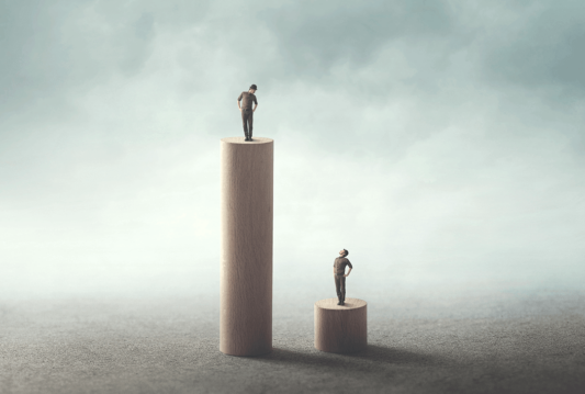 One person standing on a tall totem pole and another person standing on a short totem pole