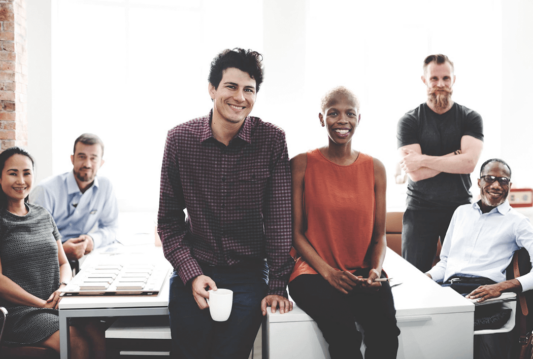 Employees in a boardroom all around a table. Two of them are standing in front of the table smiling