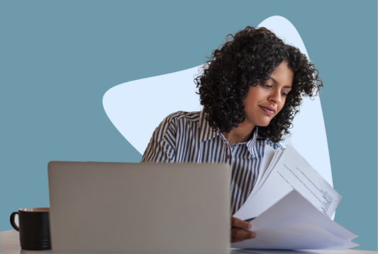 Employee sitting at desk in front of laptop and sifting through a pile of paper.