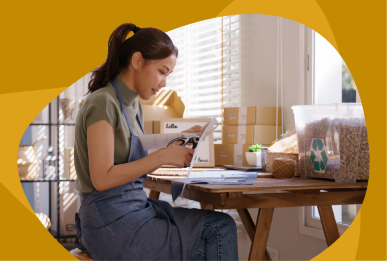 Woman with ponytail and an apron cutting paper with recycling information on it at a desk with recycle bins.