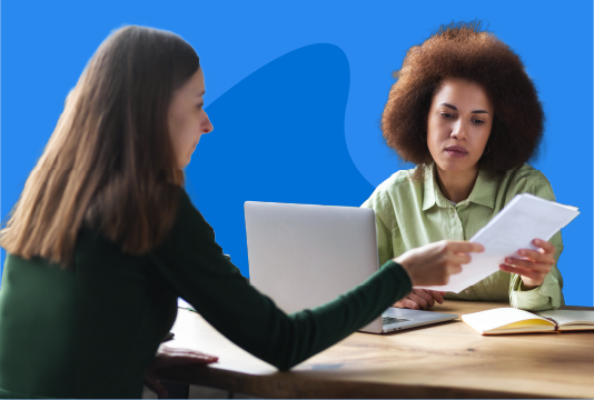 Two women are sitting across a table from each other. Both are holding the same piece of paper in their hands. The woman who is facing the camera has a laptop open. Both are staring at the paper