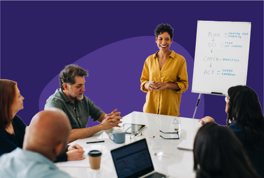 Five employees are sitting around a boardroom table. They are facing a person who is standing in front of a whiteboard presenting to the group.