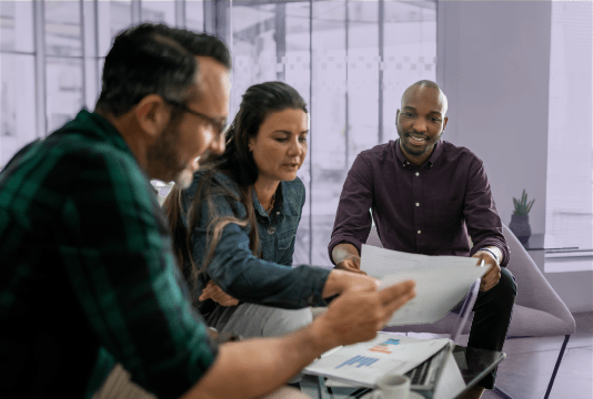 Three employees look happy as they sit around a coffee table looking at some paperwork