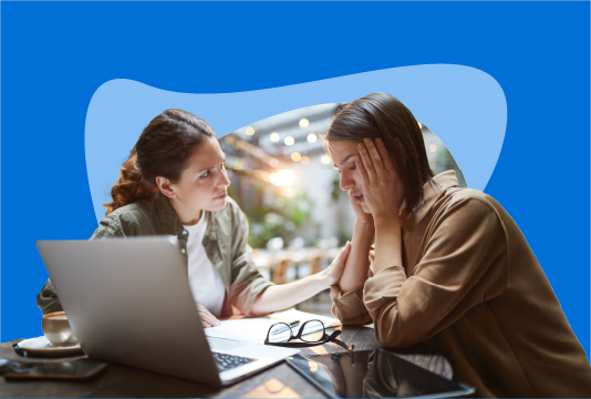 Two employees are sitting on the same side of a table. They are talking with a laptop on the table facing them. One looks distraught with her hands on her face, and the other looks concerned.