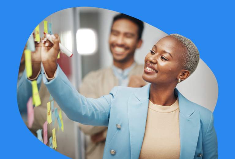A woman is writing on a clear board with a man standing behind her looking at the board. They are both smiling.
